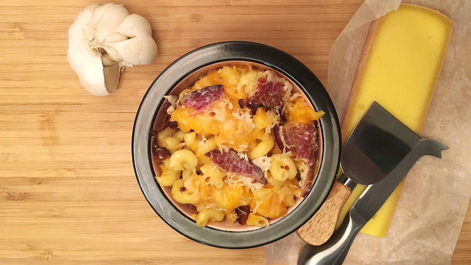 A head of garlic, a bowl of mac and cheese topped with piece of bacon, a wedge of cheese, a cheese planer and a cheese knife